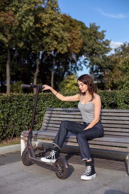 Ragazza alla moda del brunette che si siede su un banco con il suo escooter in un parco all'aperto in tempo soleggiato
