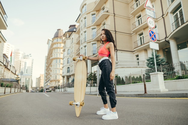 Ragazza alla moda del cavaliere in streetwear alla moda pone alla macchina fotografica con il longboard in mano