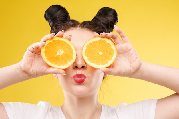 Ragazza alla moda divertente con le arance della tenuta dell'acconciatura sugli occhi.