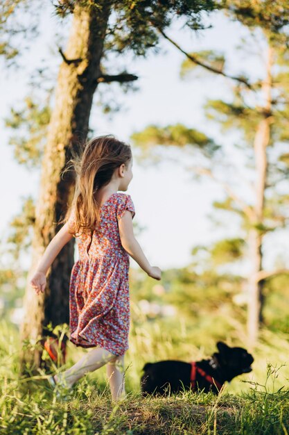 Ragazza alla moda in abito estivo elegante con un amico cane che cammina nel campo con fiori al tramonto nella natura del sole selvaggio in montagna