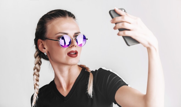 Ragazza alla moda in occhiali da sole con occhiali colorati fa selfie al telefono