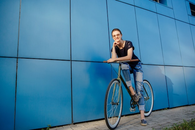 Ragazza alla moda in occhiali da sole divertirsi mentre si cammina con la bici all'aperto, indossando jeans e t-shirt.