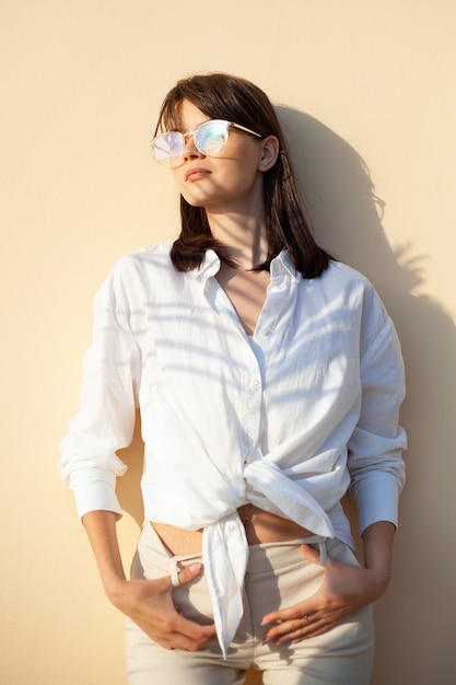 Ragazza alla moda in occhiali da sole e camicia bianca su fondo beige