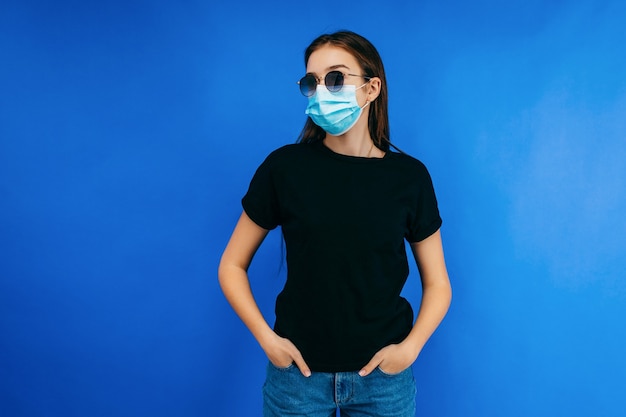 Ragazza alla moda in occhiali e maschera protettiva che indossa la maglietta nera in posa in studio sullo spazio blu