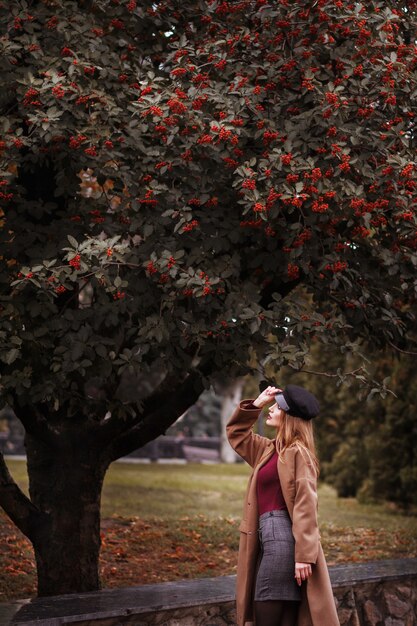 Ragazza alla moda in un cappotto vicino a un albero in un parco nel tardo autunno