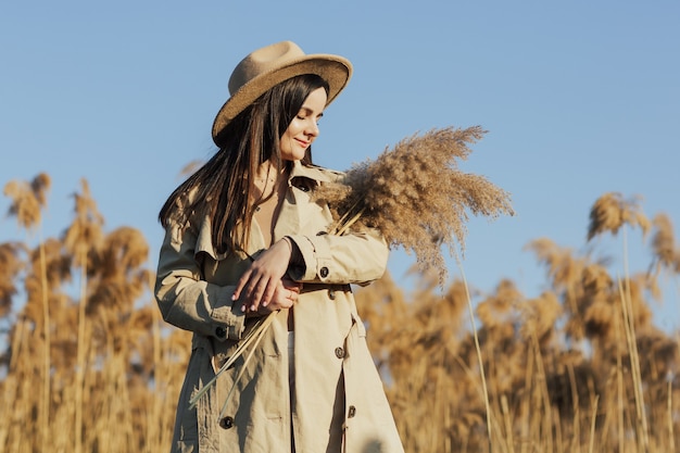 Ragazza alla moda in un trench beige e un cappello in campagna su uno sfondo di canne secche e cielo blu