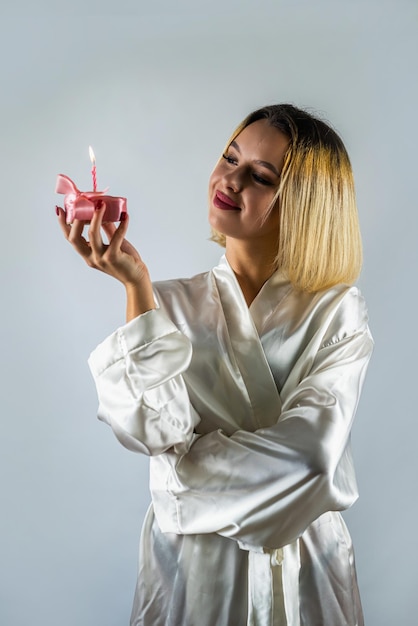 Ragazza alla moda in vestaglia bianca di seta che spegne le candeline su un pezzo di cupcake su sfondo isolato festa di compleanno