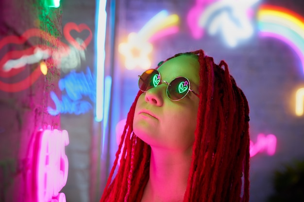 Ragazza alle luci al neon, bella donna in occhiali da sole, con capelli rosa, con trecce di dreadlocks,