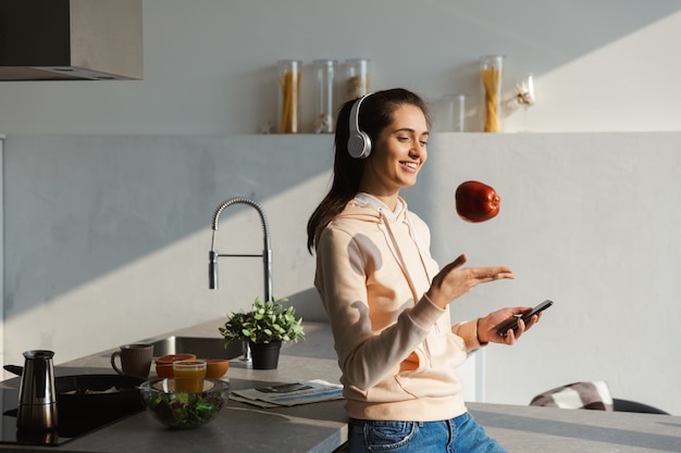 Ragazza allegra che ascolta la musica con le cuffie in cucina a casa, mangia una mela, tenendo il telefono cellulare