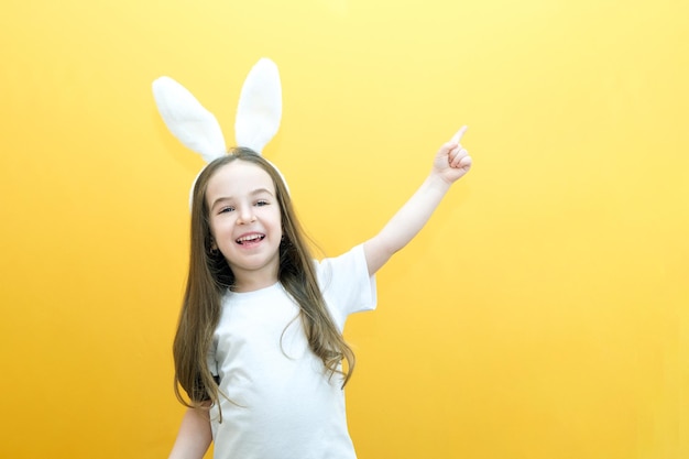 Ragazza allegra con orecchie di coniglio sulla testa su sfondo giallo Divertente bambino felice punta le dita in uno spazio vuoto copia spazio per il mockup del testo