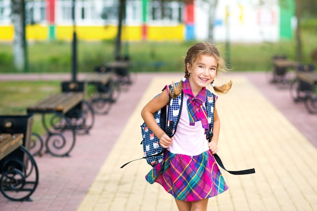 Ragazza allegra con uno zaino e in uniforme scolastica nel cortile della scuola. Ritorno a scuola, 1 settembre. Un allievo felice. Istruzione primaria, classe elementare. Ritratto di uno studente