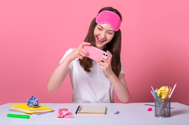 Ragazza allegra della scuola che gioca sullo smartphone invece di fare i compiti con una maglietta e una maschera per dormire