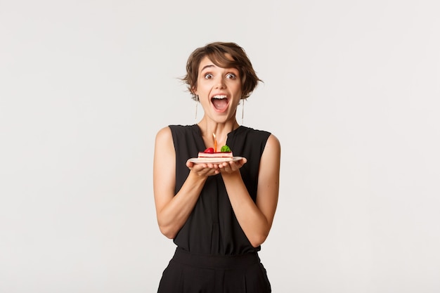 Ragazza allegra di compleanno che sembra stupita, tenendo un pezzo di torta e esprimendo il desiderio