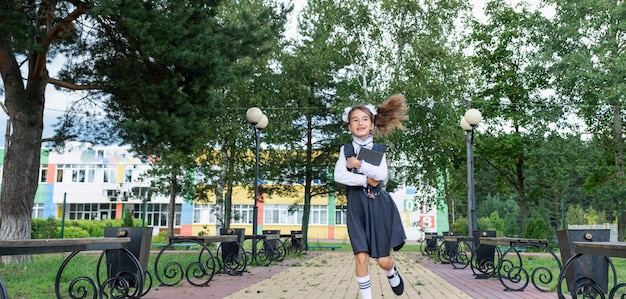 Ragazza allegra e divertente con il sorriso sdentato in uniforme scolastica con fiocchi bianchi che corre nel cortile della scuola Ritorno a scuola il 1° settembre Allievo felice con lo zaino Movimento della classe elementare dell'istruzione primaria