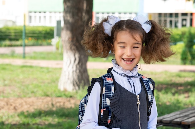 Ragazza allegra e divertente con un sorriso sdentato in uniforme scolastica con fiocchi bianchi nel cortile della scuola Ritorno a scuola 1 settembre Allievo felice con uno zaino Classe elementare di educazione primaria