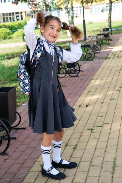 Ragazza allegra e divertente con un sorriso sdentato in uniforme scolastica con fiocchi bianchi nel cortile della scuola Ritorno a scuola 1 settembre Allievo felice con uno zaino Classe elementare di educazione primaria