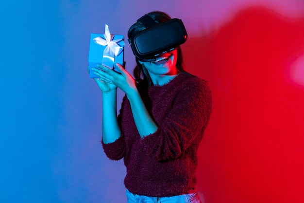 Ragazza allegra e felice che indossa occhiali per realtà virtuale VR con scatola regalo vicino all'orecchio