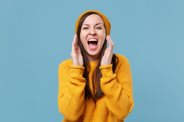 Ragazza allegra giovane donna bruna in maglione giallo e cappello in posa isolata su sfondo blu ritratto in studio. Concetto di stile di vita delle persone. Mock up spazio di copia. Urlando con il gesto della mano vicino alla bocca.