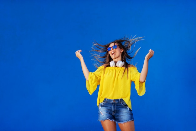 Ragazza allegra in camicia gialla che sta contro la parete blu con la cuffia avricolare intorno al suo collo.