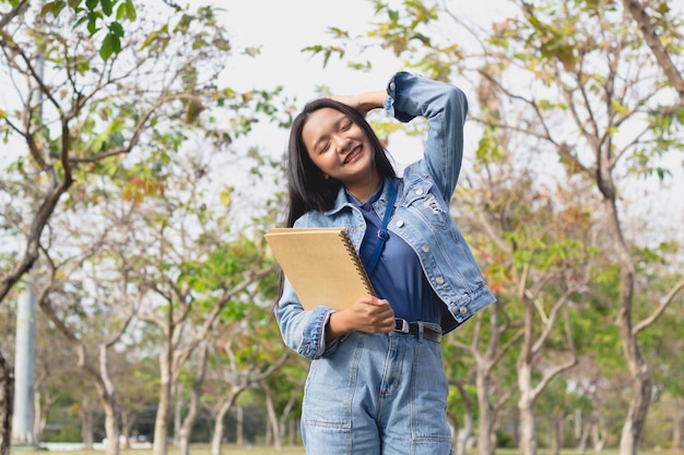 Ragazza allegra in piedi nel parco con in mano un libro e sorridente e indossa il concetto di stile di vita giacca e jeans