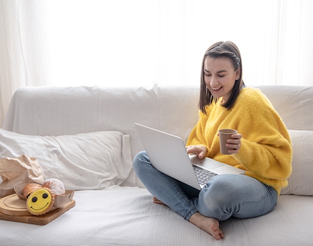 Ragazza allegra in un maglione giallo lavora a casa sul divano in remoto. Beve caffè mentre lavora.