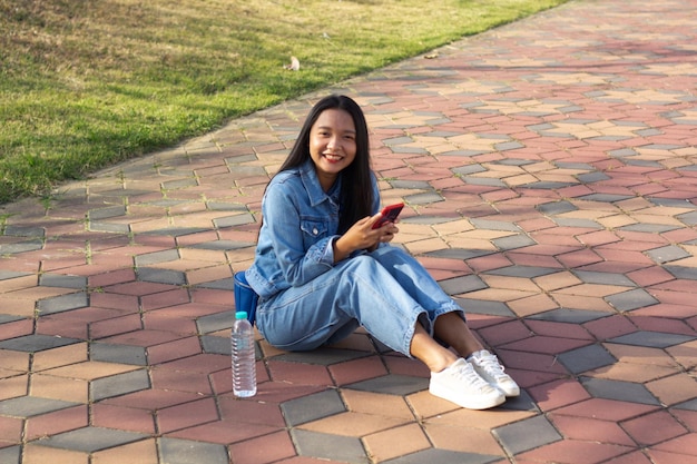 Ragazza allegra seduta al parco in città tenere il cellulare con una bottiglia d'acqua posta sul concetto di stile di vita laterale