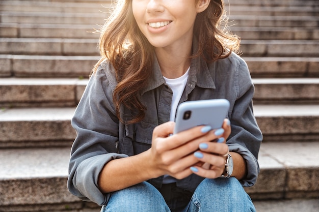 Ragazza allegra seduta sulle scale all'aperto, usando il telefono cellulare