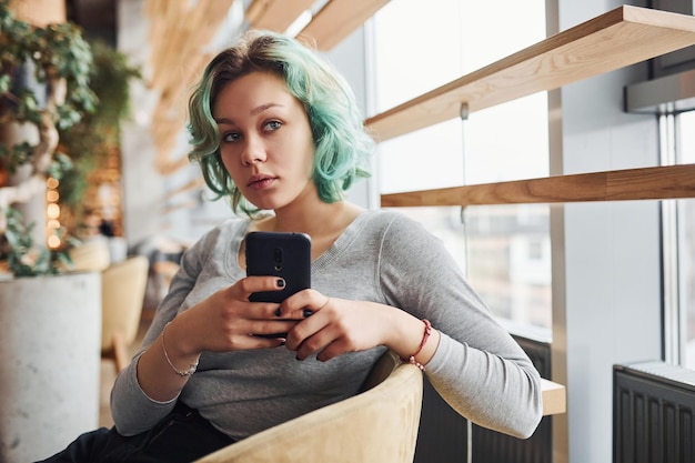 Ragazza alternativa con i capelli verdi seduta al chiuso durante il giorno con il telefono in mano.