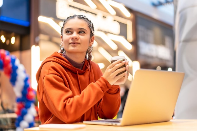 Ragazza alternativa con trecce bianche con un computer in un centro commerciale, pensierosa con un caffè caldo in mano