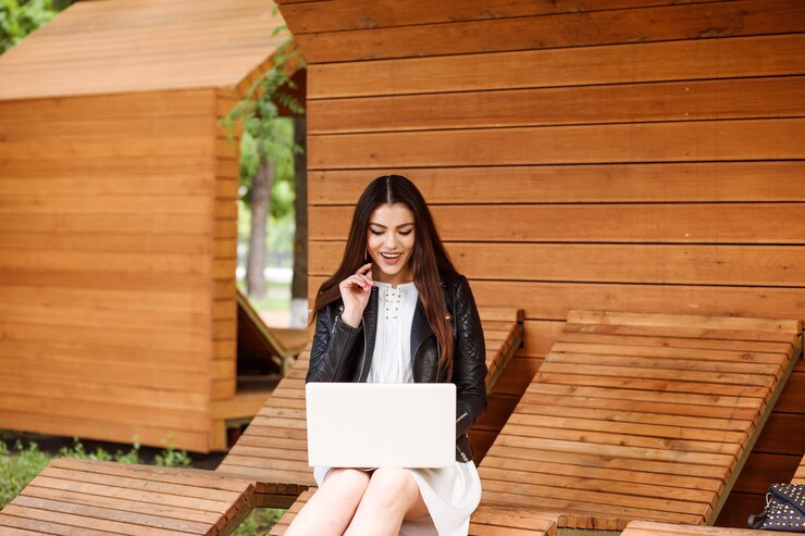 Ragazza appassionata che lavora o studia sul portatile all'aperto nel parco, seduto sulla panchina moderna. La donna sudent è vestita in abito bianco e giacca nera.