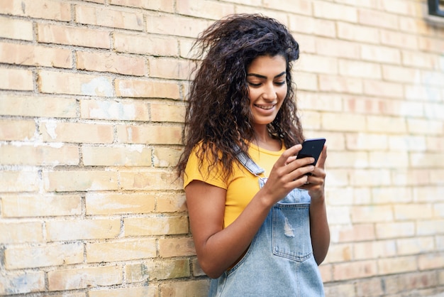 Ragazza araba felice che per mezzo dello Smart Phone sul muro di mattoni.