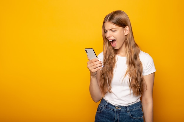 ragazza arrabbiata che grida mentre usando smartphone isolato su giallo