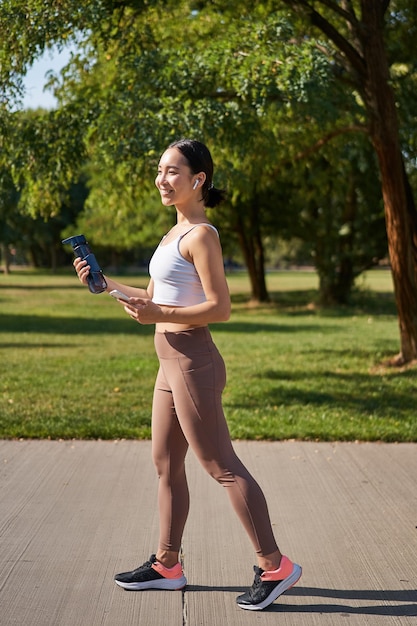 Ragazza asiatica attiva in abbigliamento fitness allenamento nel parco a piedi in abbigliamento sportivo con smartphone e wate