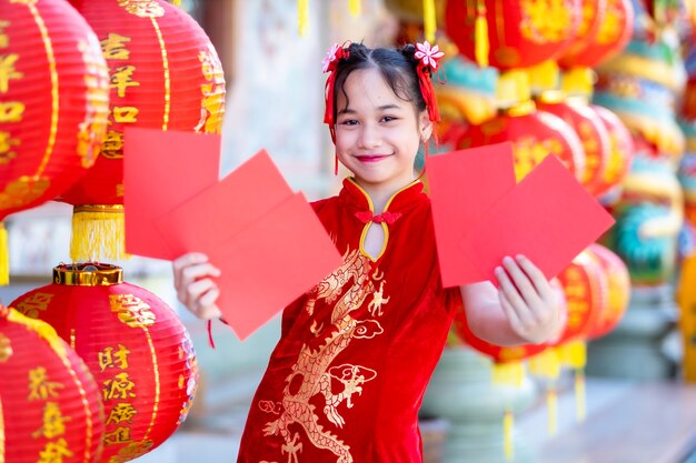 Ragazza asiatica che indossa cheongsam rosso cinese tradizionale, con buste rosse in mano e lanterne con scritto sopra il testo cinese Benedizioni È una benedizione della fortuna per il capodanno cinese