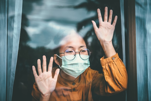 Ragazza asiatica con gli occhiali, con indosso una maschera, guardando fuori dalla finestra, è stanca di dover trattenere e curare la malattia a casa da sola. Concetto di quarantena domestica, prevenzione COVID-19