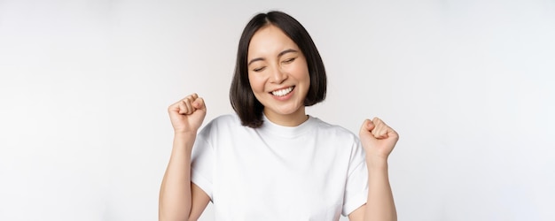 Ragazza asiatica danzante che celebra sentirsi felice e ottimista sorridente ampiamente in piedi su sfondo bianco studio