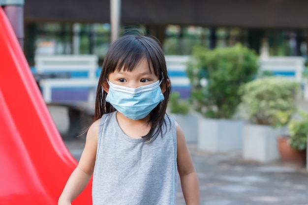 Ragazza asiatica del bambino che sorride e maschera del tessuto da portare Lei che gioca con il giocattolo della barra del cursore al parco giochi