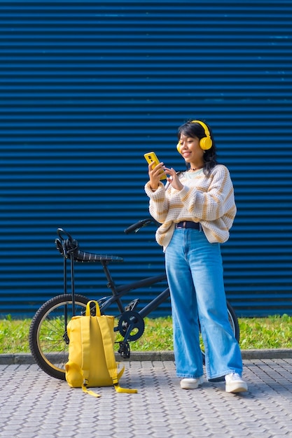 Ragazza asiatica del college che ascolta musica con le cuffie gialle su uno sfondo blu del college