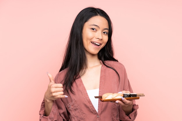 Ragazza asiatica dell'adolescente che mangia sushi isolato sulla parete rosa che fa gesto del telefono