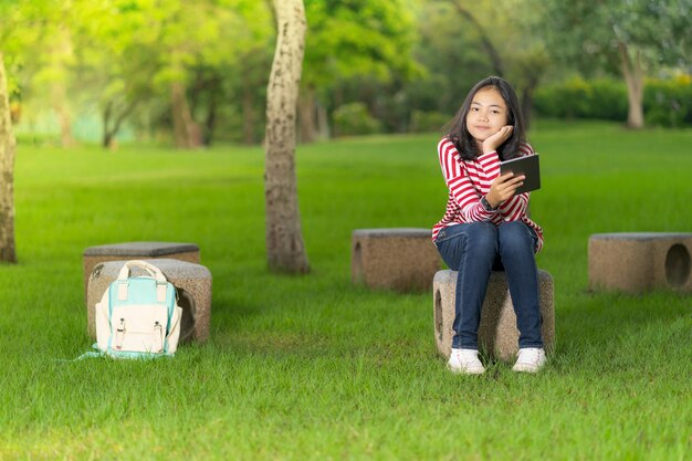 Ragazza asiatica dello studente con una compressa digitale nel parco della scuola in un giorno di estate soleggiato