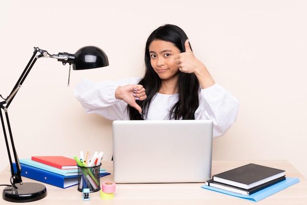 Ragazza asiatica dello studente in un posto di lavoro con un computer portatile sulla parete beige che fa segno buono-cattivo. Indeciso tra sì o no