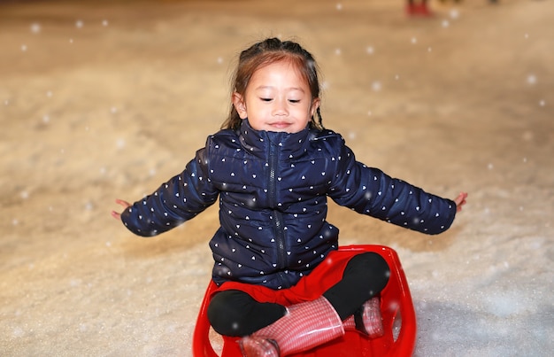 Ragazza asiatica felice del bambino che gioca sul vassoio di plastica rosso in neve ad orario invernale