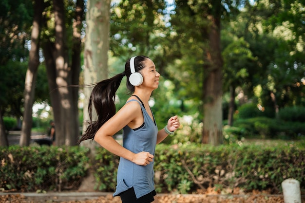 Ragazza asiatica felice dell'atleta che corre nel parco mentre sta ascoltando la musica sulle cuffie bianche