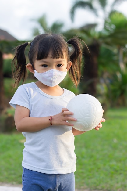 Ragazza asiatica giocosa felice del bambino che porta maschera facciale del tessuto. Gioca con la palla nel parco giochi del parco verde.