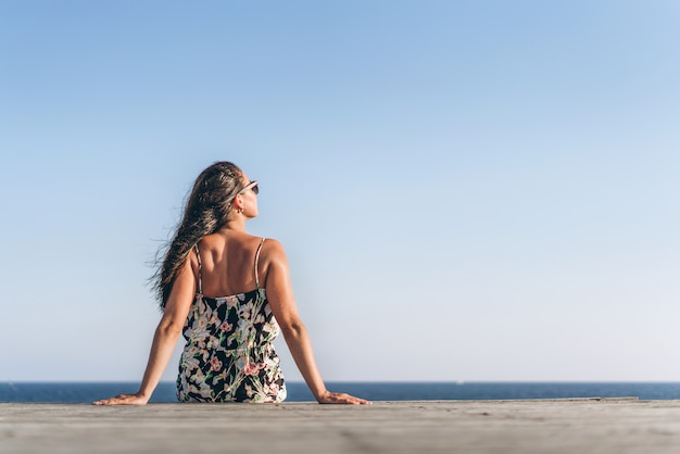 Ragazza asiatica graziosa della pentola in vestito che si siede sul pilastro vicino al mare.