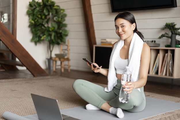 Ragazza asiatica in abbigliamento fitness seduta a casa sul tappetino che riposa dopo l'allenamento controllando il cellulare...