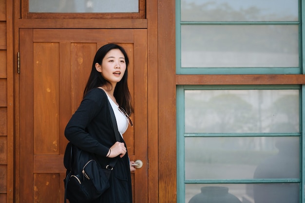 ragazza asiatica in maglietta bianca con zaino aprire la vecchia porta di legno della casa del trono. donna affascinante che guarda con curiosità mentre gira la testa e trova qualcuno che la chiama. femmina che cammina in casa.