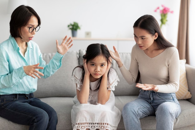 Ragazza asiatica infelice che copre le orecchie mentre la madre e la nonna arrabbiate la rimproverano a casa della famiglia