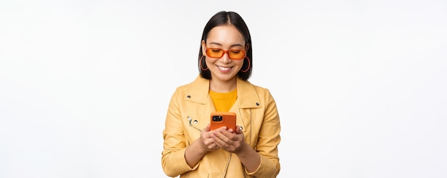 Ragazza asiatica moderna in occhiali da sole utilizzando il suo telefono cellulare sorridente e guardando felice in posa su sfondo bianco
