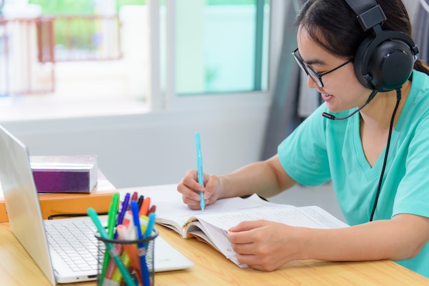 Ragazza asiatica studentessa adolescente con occhiali cuffie seduta sorriso felice guardando scrivere note al libro computer portatile sul tavolo apprendimento online studio istruzione dalla classe dell'università a casa